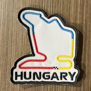 May include: A white embroidered patch featuring the Hungaroring race track layout outlined in blue, red, and yellow thread. The word HUNGARY is stitched in bold black capital letters at the bottom, all enclosed by a thick black border.