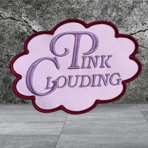 Puede incluir: Un parche bordado con forma de nube que presenta un fondo rosa claro con un borde granate oscuro. El texto "PINK CLOUDING" est&aacute; cosido en una fuente cursiva de color p&uacute;rpura en el centro del parche, creando un contraste visual distintivo.