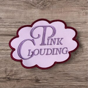 Puede incluir: Un parche bordado con forma de nube con fondo rosa claro y borde granate oscuro. El texto PINK CLOUDING est&aacute; bordado con hilo morado en el centro en una fuente serif decorativa con una P y una C grandes.