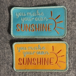 May include: Two rectangular embroidered patches. One is turquoise with the words "you make your own SUNSHINE" in white and orange, with a sun graphic. The other is orange with the same text and sun graphic.