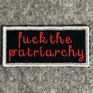 May include: A black patch with white stitched edging. The patch features the phrase "fuck the patriarchy" embroidered in red cursive lettering. The patch is rectangular in shape.