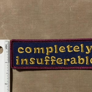 Peut inclure: Un &eacute;cusson rectangulaire brod&eacute; avec un fond bleu fonc&eacute; et une bordure rouge fonc&eacute;. Le texte "completely insufferable" est cousu en fil dor&eacute;. Une r&egrave;gle blanche est plac&eacute;e &agrave; gauche, montrant que l'&eacute;cusson mesure 7,6 cm de large.