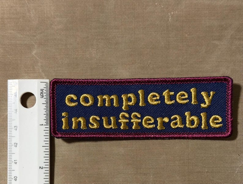 Peut inclure: Un &eacute;cusson rectangulaire brod&eacute; avec un fond bleu fonc&eacute; et une bordure rouge fonc&eacute;. Le texte "completely insufferable" est cousu en fil dor&eacute;. Une r&egrave;gle blanche est plac&eacute;e &agrave; gauche, montrant que l'&eacute;cusson mesure 7,6 cm de large.