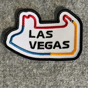 May include: A white embroidered patch with a black outline depicting the Las Vegas Motor Speedway race track. The track is outlined in red, blue, and yellow. The words "LAS VEGAS" are embroidered in black.