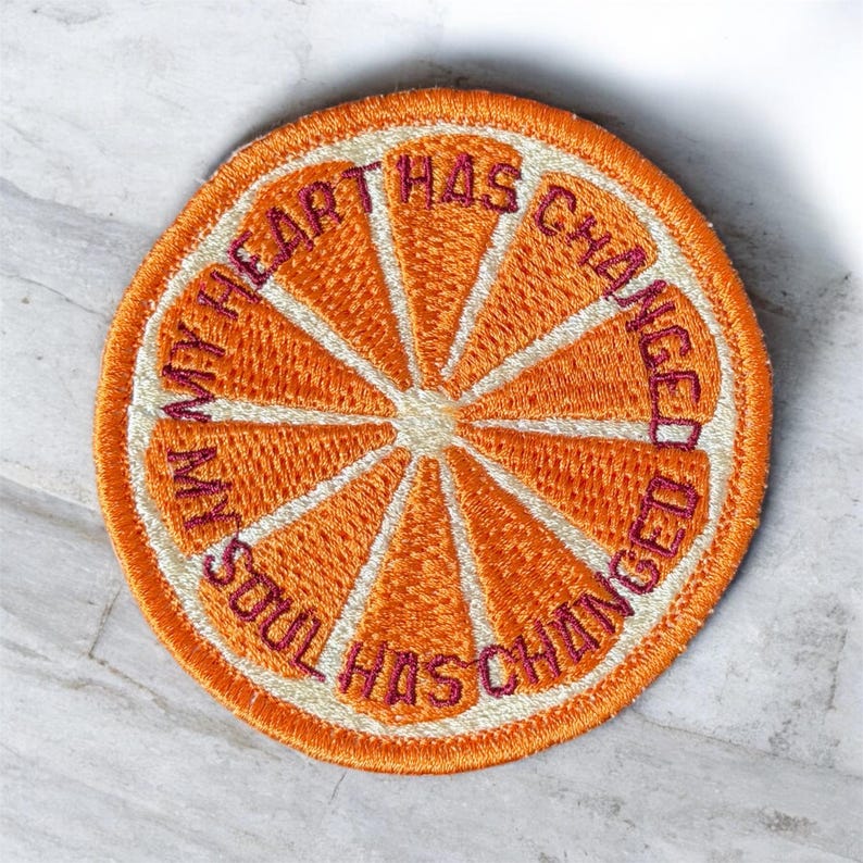 May include: Embroidered patch in the shape of an orange slice. The patch is orange with white and purple accents. The text "MY HEART HAS CHANGED, MY SOUL HAS CHANGED" is embroidered in purple around the edge of the orange slice.