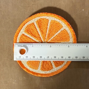 May include: Embroidered orange slice patch, approximately 3 inches in diameter. The patch features a bright orange color with white segments and a white border. The design is a detailed representation of a citrus fruit.