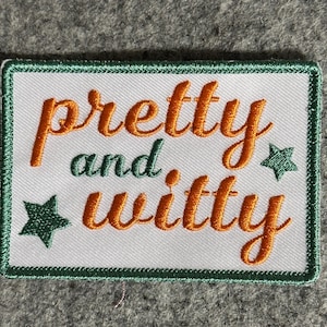 May include: A rectangular patch with the words "pretty and witty" embroidered in orange script on a white background. The patch has a green border and two green star accents.