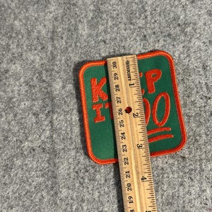 May include: A green and orange embroidered patch with the words "KEEP IT 100" in orange. The patch is rectangular with rounded corners and is approximately 3 inches in length. The patch is on a gray surface.