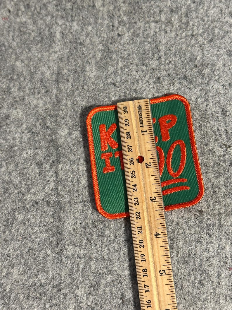 May include: A green and orange embroidered patch with the words "KEEP IT 100" in orange. The patch is rectangular with rounded corners and is approximately 3 inches in length. The patch is on a gray surface.