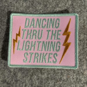 Könnte beinhalten: Rechteckiger, rosa Aufnäher mit dem Text "DANCING THRU THE LIGHTNING STRIKES" in Hellgrün. Zwei goldene Blitze flankieren den Text. Der Aufnäher hat einen hellgrünen Rand.