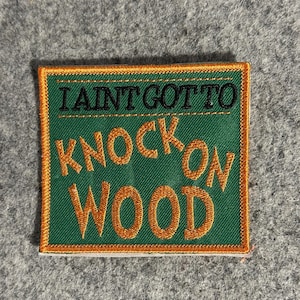 May include: Square embroidered patch with the text "I AINT GOT TO KNOCK ON WOOD" in black and gold thread on a green background. The patch has a gold border.