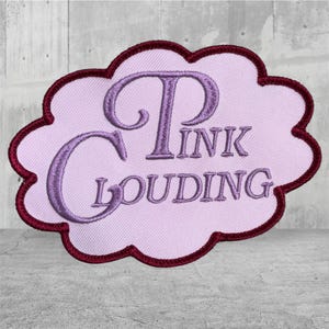 May include: A cloud-shaped embroidered patch featuring a light pink background with a dark maroon border. The text PINK CLOUDING is stitched in a purple, stylized serif font across the center of the patch, creating a distinct visual contrast.