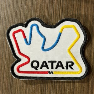 May include: A white embroidered patch featuring a black border and a stylized race track outline in red, blue, and yellow thread. The word QATAR is stitched in bold black capital letters below the track design on the white fabric background.