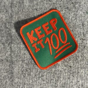May include: A square patch with a green background and orange trim. The patch features the words "KEEP IT 100" in orange, with the "100" stylized with the hundred points emoji.