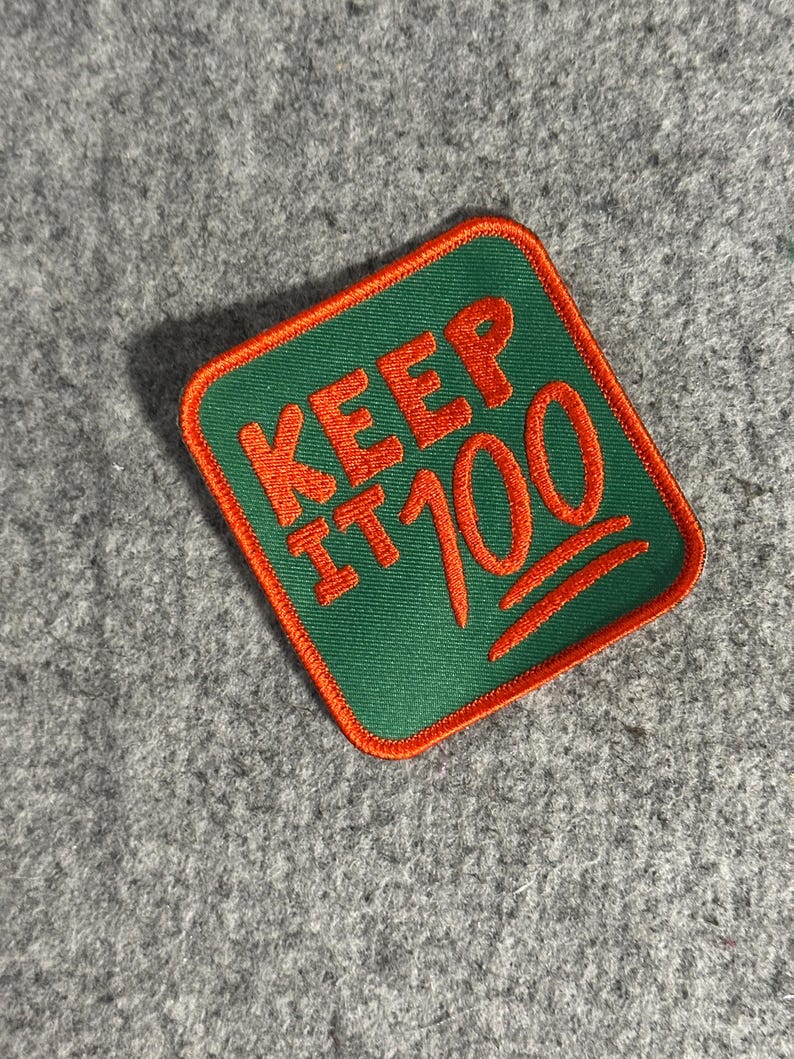 May include: A square patch with a green background and orange trim. The patch features the words "KEEP IT 100" in orange, with the "100" stylized with the hundred points emoji.