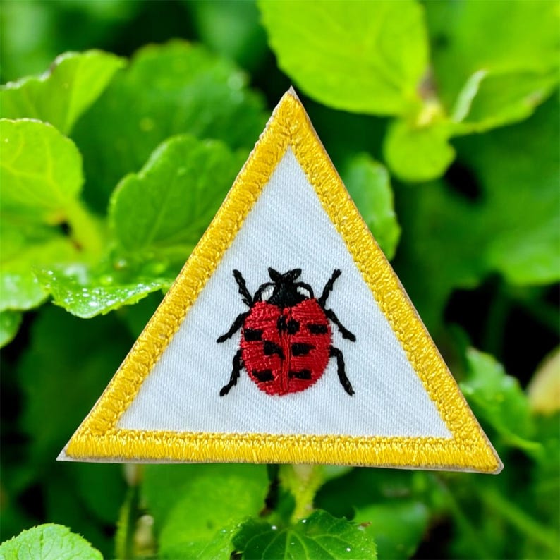 May include: A triangular patch featuring a white background, a yellow embroidered border, and a red ladybird design with black spots and legs. Suitable for sewing onto clothing or bags.