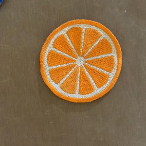 May include: Embroidered patch featuring a vibrant orange slice design. The patch has a bright orange center with white segments and a white border. This circular patch is suitable for clothing or accessories.