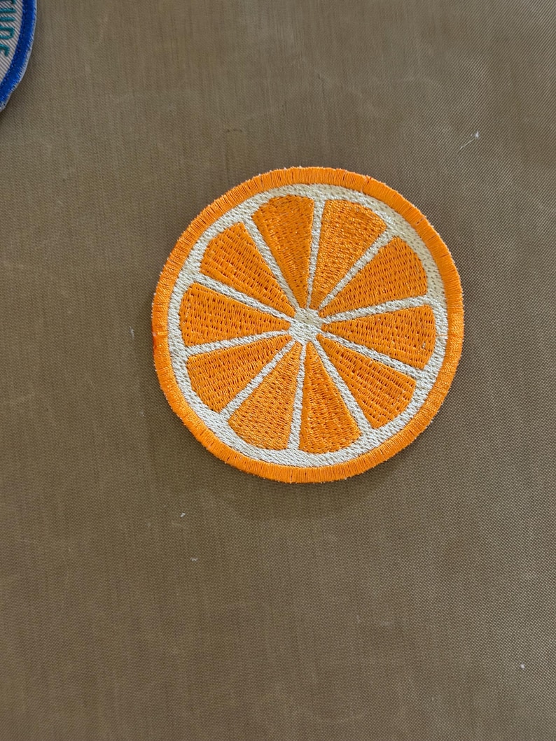 May include: Embroidered patch featuring a vibrant orange slice design. The patch has a bright orange center with white segments and a white border. This circular patch is suitable for clothing or accessories.