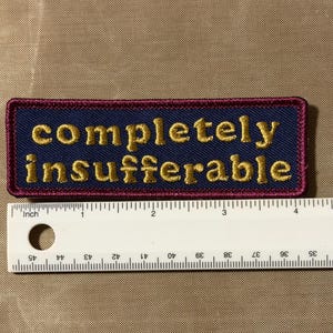 Peut inclure: Un &eacute;cusson rectangulaire brod&eacute; avec un fond bleu fonc&eacute; et une bordure rouge fonc&eacute;. Le texte "completely insufferable" est cousu en fil dor&eacute;. Une r&egrave;gle blanche est plac&eacute;e en dessous, montrant que l'&eacute;cusson mesure 10,2 cm de large.