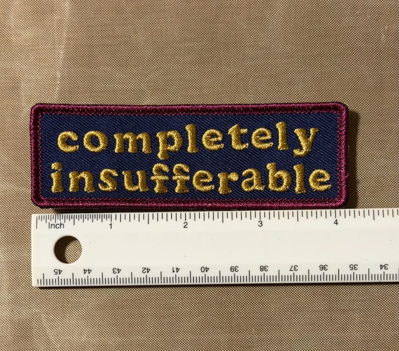 Peut inclure: Un &eacute;cusson rectangulaire brod&eacute; avec un fond bleu fonc&eacute; et une bordure rouge fonc&eacute;. Le texte "completely insufferable" est cousu en fil dor&eacute;. Une r&egrave;gle blanche est plac&eacute;e en dessous, montrant que l'&eacute;cusson mesure 10,2 cm de large.