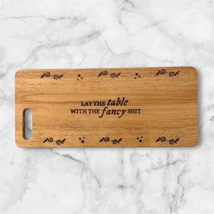 May include: Wooden rectangular serving board with a handle. The board has decorative leaf and star patterns and the text "LAY THE table WITH THE fancy shit" engraved on it. The board is made of light brown wood.