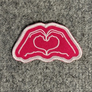 May include: A pink embroidered patch featuring a heart shape formed by two hands. The patch has a white border and is set against a gray background. The design is a symbol of love and affection.