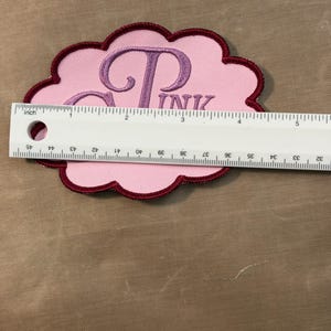 Puede incluir: Un parche bordado con forma de nube con un centro rosa claro y un borde granate oscuro. El parche presenta el texto bordado en violeta "PINK" y una letra "P" grande. Una regla blanca est&aacute; colocada sobre el parche, mostrando 12,7 cm de ancho.