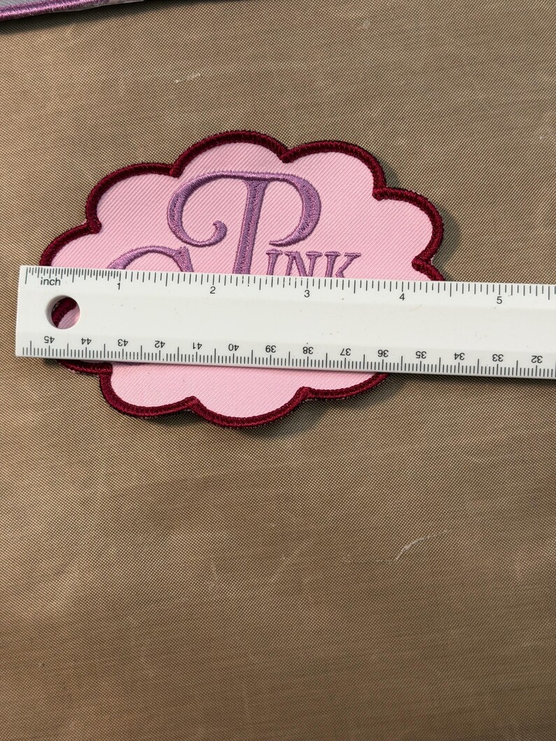 Puede incluir: Un parche bordado con forma de nube con un centro rosa claro y un borde granate oscuro. El parche presenta el texto bordado en violeta "PINK" y una letra "P" grande. Una regla blanca est&aacute; colocada sobre el parche, mostrando 12,7 cm de ancho.
