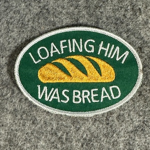 May include: An oval patch with a green background and white border. The patch features the text "LOAFING HIM WAS BREAD" in white, surrounding a golden-yellow illustration of a loaf of bread.