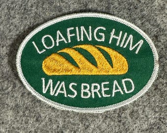Peut inclure: Un écusson ovale avec un fond vert et une bordure blanche. L'écusson présente le texte "LOAFING HIM WAS BREAD" en blanc, entourant une illustration jaune doré d'une miche de pain.
