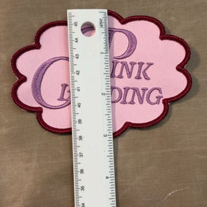 Puede incluir: Un parche bordado en forma de nube con fondo rosa claro y borde rojo oscuro. El parche presenta texto bordado en color p&uacute;rpura que dice "DINK G DING". Una regla blanca est&aacute; colocada verticalmente sobre el parche, mostrando un ancho de 7,6 cm.