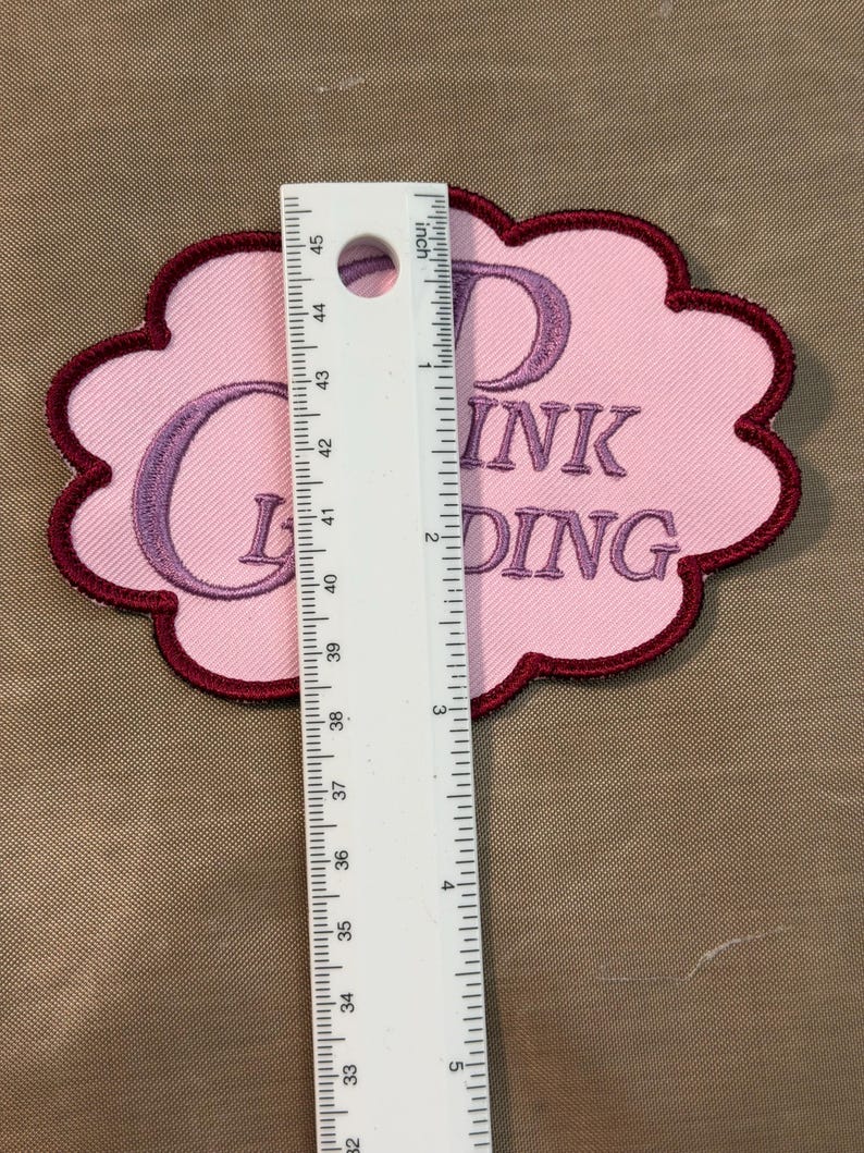 Puede incluir: Un parche bordado en forma de nube con fondo rosa claro y borde rojo oscuro. El parche presenta texto bordado en color p&uacute;rpura que dice "DINK G DING". Una regla blanca est&aacute; colocada verticalmente sobre el parche, mostrando un ancho de 7,6 cm.