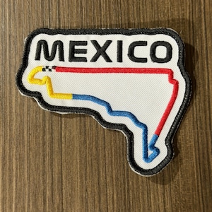 May include: A white embroidered patch with a black border featuring the word MEXICO in black capital letters above a stylized outline of a racetrack. The track outline is colored with yellow, blue, and red segments and includes a small checkered flag.