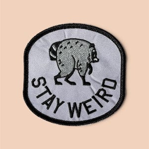 May include: A circular embroidered patch with a light gray background and a thick black border. The patch features a gray raccoon illustration in the center and the black embroidered text "STAY WEIRD" curved along the bottom edge.
