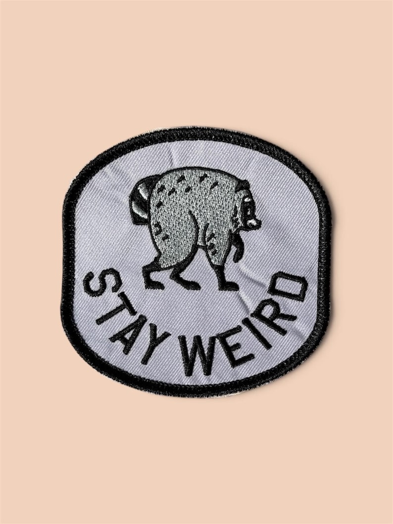 May include: A circular embroidered patch with a light gray background and a thick black border. The patch features a gray raccoon illustration in the center and the black embroidered text "STAY WEIRD" curved along the bottom edge.