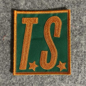 Puede incluir: Parche bordado verde con las letras doradas "TS", enmarcado por un borde dorado. Dos estrellas doradas en la parte inferior. El parche es cuadrado y tiene una apariencia texturizada.