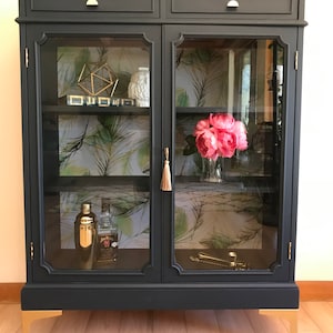 May include: A black cabinet with glass doors and gold legs. The cabinet has two shelves and is decorated with a tropical print wallpaper. There are various items on the shelves, including a cocktail shaker, a bottle of liquor, and a vase with pink flowers.
