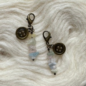 May include: Two antique gold-toned stitch markers with a button charm and a strand of pastel-colored gemstone beads.