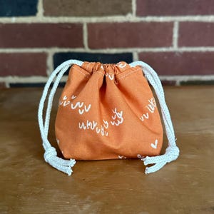May include: A small orange drawstring bag with white doodle-like patterns and a white heart. The bag has white drawstrings.