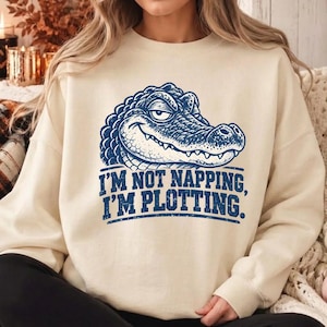 May include: A cream-colored crewneck sweatshirt featuring a blue graphic of an alligator with a sly expression. Below the alligator, the text reads, "I'M NOT NAPPING, I'M PLOTTING." The garment has long sleeves and a ribbed collar.