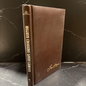 May include: A brown hardcover book titled "Mojave Crossing" by Louis L'Amour. The book is part of "The Louis L'Amour Collection." The author's signature is in gold on the cover. The book has a textured appearance.
