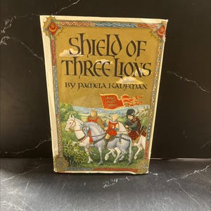 May include: A hardcover book titled "Shield of Three Lions" by Pamela Kaufman. The cover features an illustration of three figures on horseback, with a red flag bearing a crest. The book has a gold and cream-colored border.