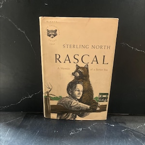 May include: Vintage hardcover book titled "Rascal" by Sterling North. The cover features an illustration of a boy with a raccoon, with the title in large, bold letters. The book is a memoir and has a "Dutton Animal Book Award" seal.