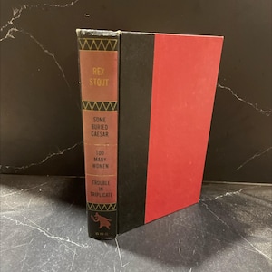 May include: A hardcover book by Rex Stout, featuring a red spine with the titles "Some Buried Caesar", "Too Many Women", and "Trouble in Triplicate". The book has a black spine and a red cover.