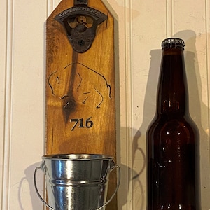 May include: Wooden wall mounted bottle opener with a metal bottle opener, a small metal bucket, and a brown glass bottle. The bottle opener has the words "Open Here" engraved on it. The wooden board has a buffalo silhouette and the number "716" engraved on it.