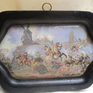 May include: Black, hexagonal tray featuring a historical battle scene print. The print shows soldiers on horseback and on foot, with a burning building and a windmill in the background. Includes a small metal ring for hanging.