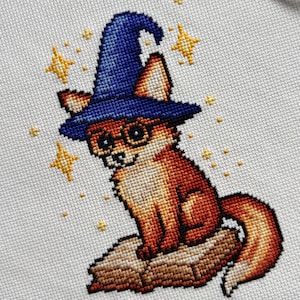 May include: Cross-stitch of a fox wearing a blue wizard hat and glasses, sitting on a log. The fox is orange and white, with yellow stars around it. The background is a light beige fabric.