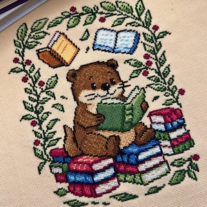 May include: Cross-stitch of a brown otter reading a green book, surrounded by a green and red floral arch. The otter sits on a stack of colorful books. Two books float above the otter. The background is a beige fabric.