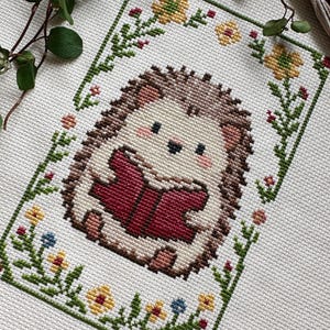 May include: Cross-stitch of a cute hedgehog reading a book, surrounded by a floral border. The hedgehog has brown quills, a cream-colored body, and is holding a red book. The border features green leaves, yellow, red, and blue flowers.