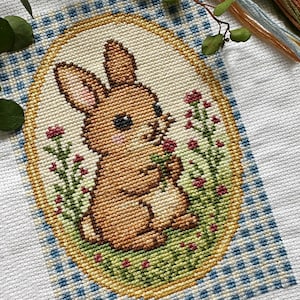 Puede incluir: Un diseño de punto de cruz con un conejito marrón sosteniendo flores, enmarcado en un óvalo. El marco está rodeado por un patrón de cuadros azules y blancos sobre un fondo blanco. Se ve vegetación y hilo de bordar.